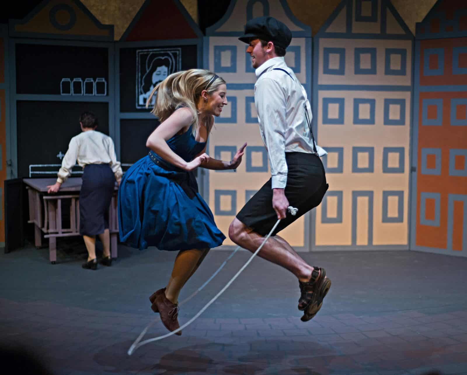 a young blond female wears a blue dress and jumps rope with a young man in white shirt and breeches