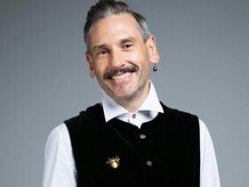 A person wearing a white button up, black vest, a bee broach and a dark opal earring poses smiling.