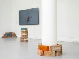 stacked glass and red bricks. TV instillation in the background featuring the artist lifting bricks.