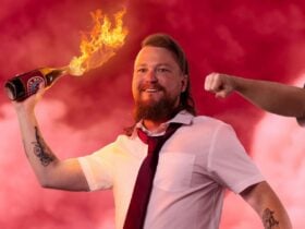 Comedian Brett Blake holding a flaming beer bottle in front of a cloudy pink background