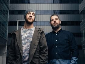 Two men standing in front of an office building. One wears a jacket and beanie, the other a shirt