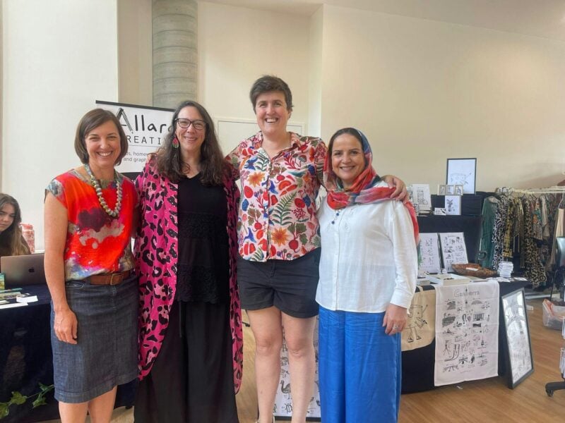 Four women stand before market stalls at the For Purpose Markets