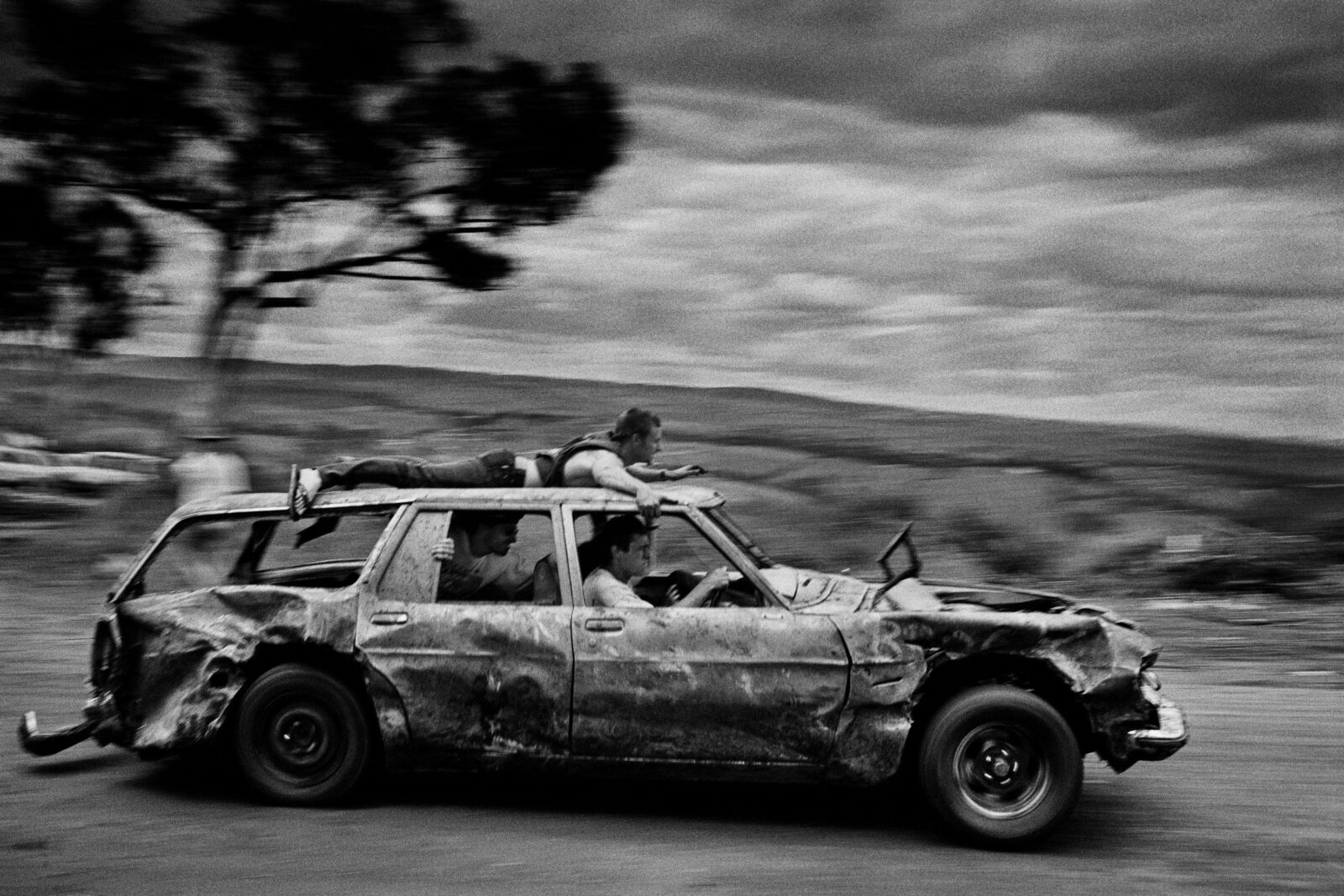 black & white image of a banged-up car speeding past with passengers and one man on the car roof