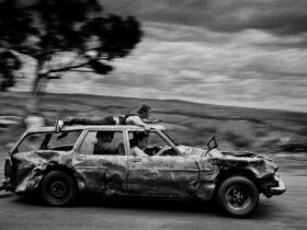 black & white image of a banged-up car speeding past with passengers and one man on the car roof