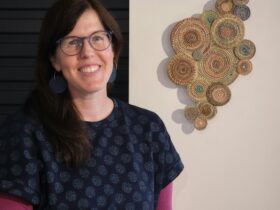 Sally Holliday stands beside her woven art handing on the wall behind her.