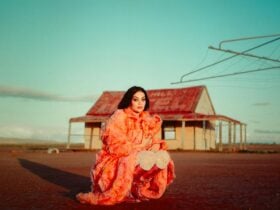 Gamilaraay musician Thelma Plum, wearing a coral-coloured feather coat, kneels on Wilyakali Country.