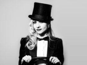 Nikki Nouveau in top hat, bow tie and tails, straddles a chair, facing the viewer.