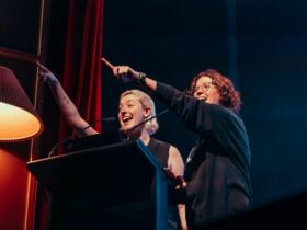 Hosts Harriet Gillies and Xanthe Dobbie on-stage behind a lectern, joyously pointing at audience.