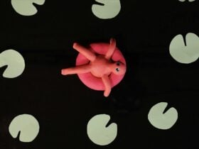 A video still image of a pink clay character sitting in a pool float surrounded by green lily-ads