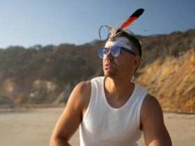 Mitch Tambo sits on a beach with a rocky coastline looking out to sea.