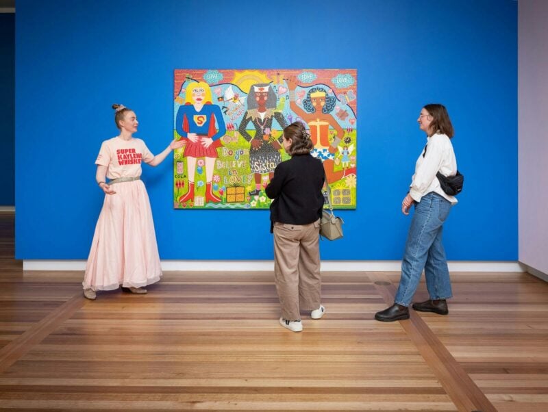 A guide in a pink outfit speaks to two visitors about a colourful portrait.