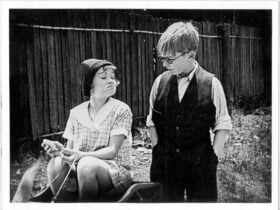 Black and white image of girl smirking and sitting down and boy with glasses looking down at her