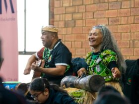 2 performers play music for an audience, one playing a guitar and one playing percussion instrument