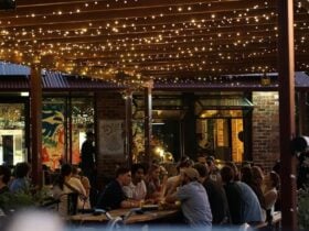 People sitting along outdoor tables in the evening time, underneath fairy lights