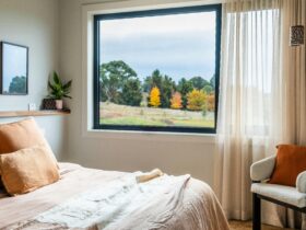 a bed covered in orange linen with an open window and blinds.