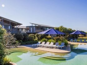 Two beach houses overlooking a shallow pool surrounded by white sunlounges and blue umbrellas