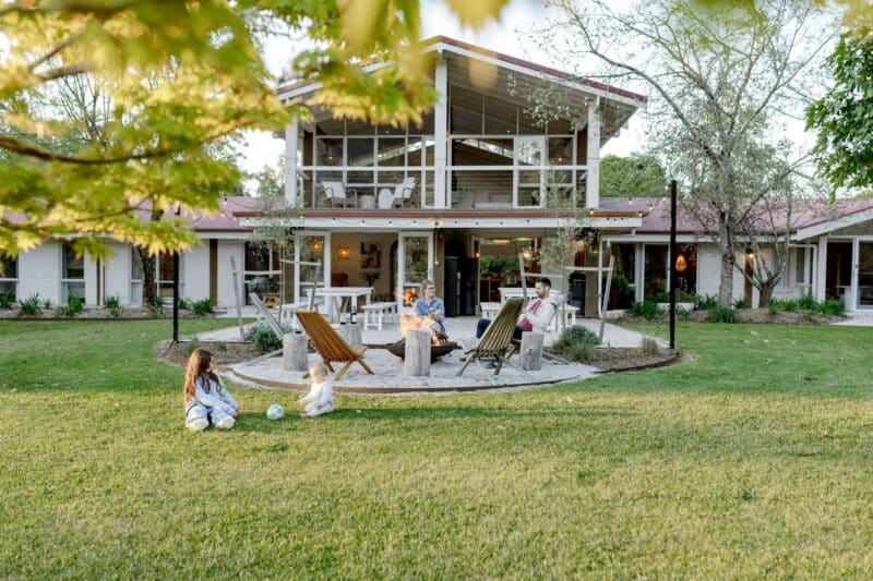 Image of front of home. GUests are sitting around firepit enjoying the bush view. Baby and Girl play