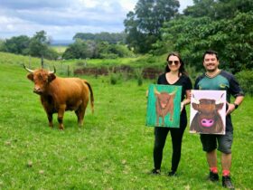 2 women holding their highland cow painting in the paddock with a highland cow
