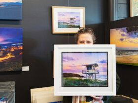 photographer behind local photo in white frame with other local photos on the wall behind.