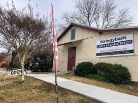 Left front view of cream fibro building, iron roof and red door with Open sign and flag at front