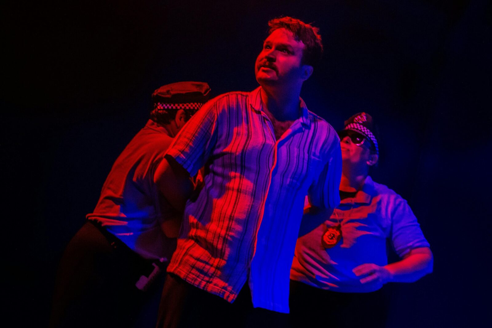A man in red and blue lighting, his arms held behind him by two police officers.