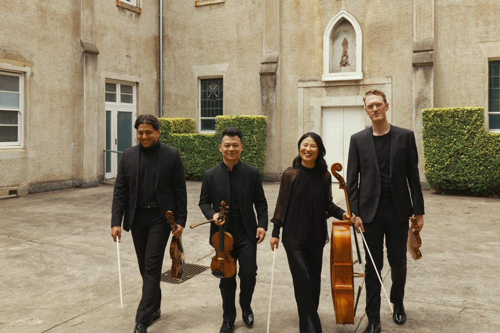 Four musicians walk together holding their instruments with a historic convent as the background