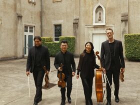 Four musicians walk together holding their instruments with a historic convent as the background