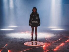 A person in a dark hoodie stands inside a glowing pentagram on a dimly lit floor