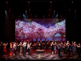 A large group of performers stand on a theater stage with a backdrop of cherry blossom trees