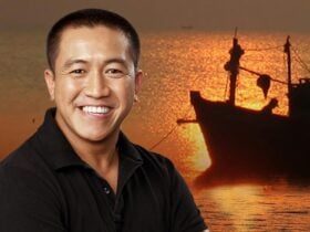 Smiling man in a black shirt stands in front of a scenic sunset with a silhouette of a boat