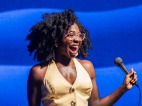 A woman with curly hair and glasses smiles widely while holding a microphone
