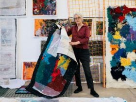 Elizabeth casually holding up a textile work, wearing a red shirt and black pants