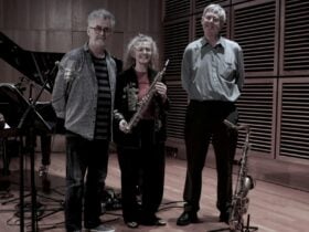 Three people stand in a recording studio with wood-paneled walls; one holds a clarinet