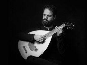 Hamid holds a classical guitar whilst performing. Black backdrop.