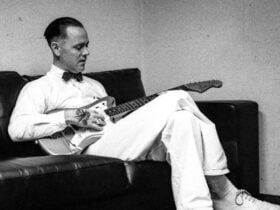 C.W. Stoneking sits on a couch in an all white outfit holding a banjo