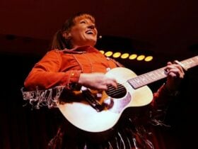 A smiling woman in a fringed red jacket plays an acoustic guitar onstage under warm yellow lighting,