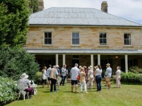 People gather out the front of a historic two-storey sandstone cottage