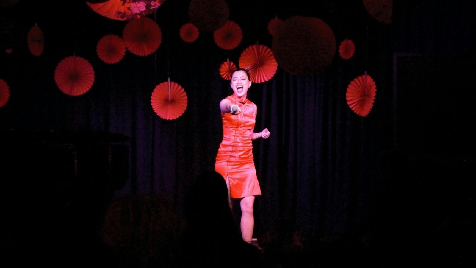 A woman in a vibrant red dress sings passionately on stage, surrounded by red paper decorations, aga