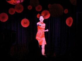 A woman in a vibrant red dress sings passionately on stage, surrounded by red paper decorations, aga