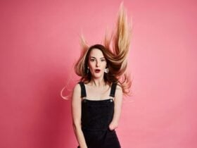 MC Madeline Stewart in front of a pink background wearing black overalls