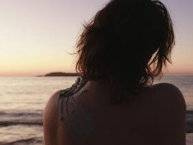 An image of a woman, from the back, rubbing her bare shoulder with white ochre in front of an ocean