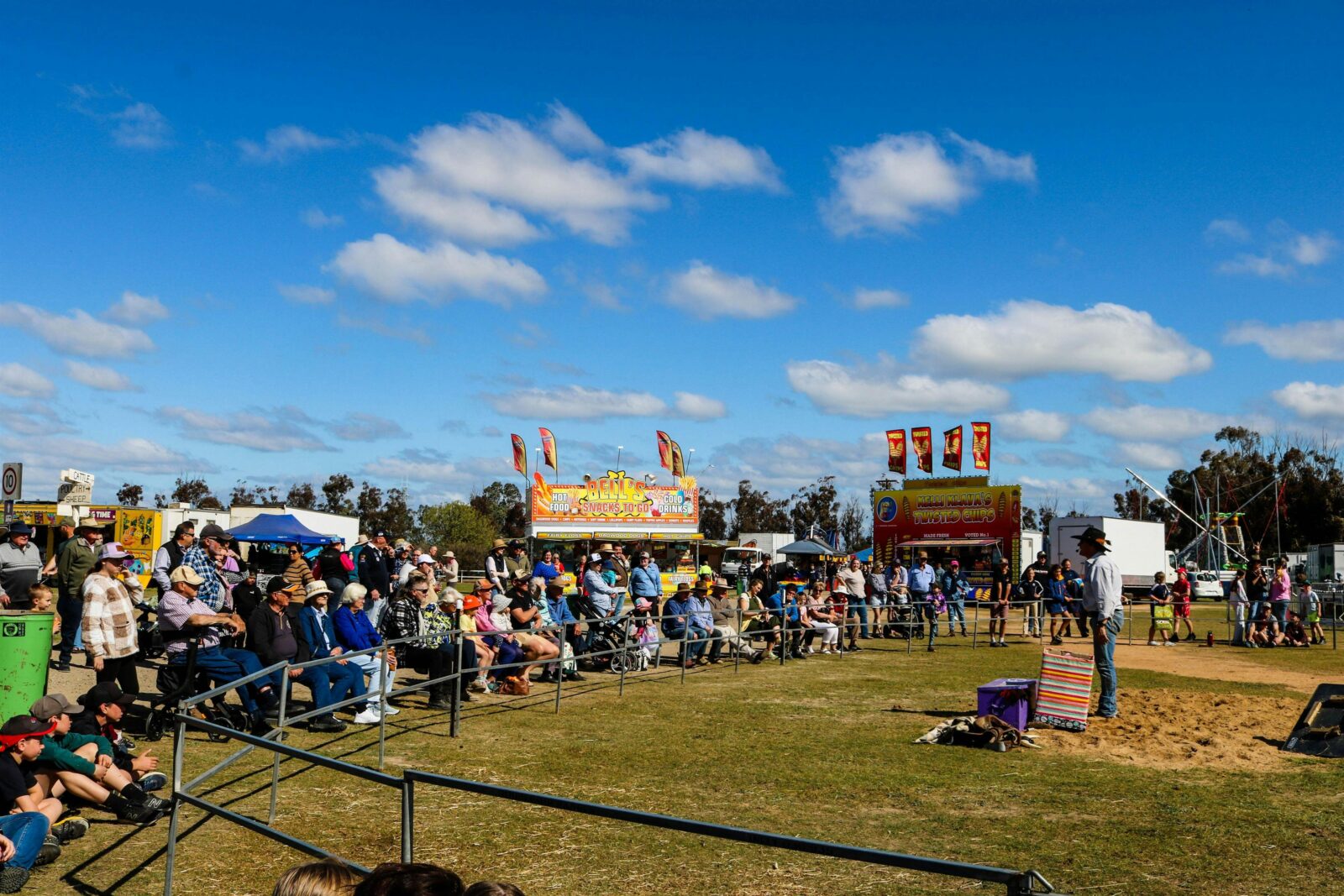 Large crowd sitting around a fence watching a stockman perform, with sideshows in background.