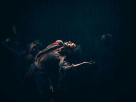 A person poses dramatically under stage lighting, arching their back and extending one arm