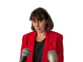 A woman with shoulder-length dark hair and a red blazer stands in front of a white backdrop