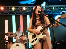 A woman plays electric guitar and sings on stage, with a drummer performing behind her.