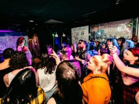 A diverse group of people crowd around a stage as a drag queen performs a lipsync