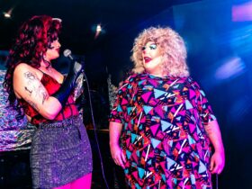 Two drag queens stand together on stand - one holds a microphone and is talking to the other