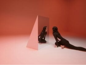 Image of a woman dressed in black looking into a mirrored object in a red hued room.