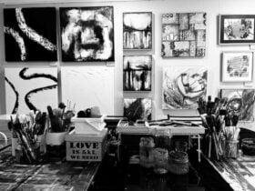 A black and white photo of an art studio table with a variety of paintings on the wall