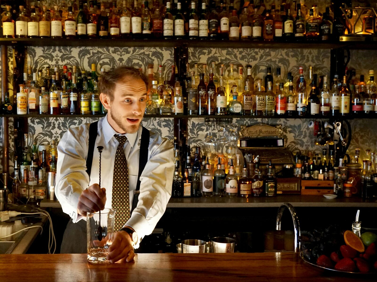 A performing bartender crafts a cocktail behind the bar while talking to the audience